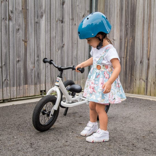 Child wearing a blue CoConuts helmet and floral dress standing next to a white Trybike balance bike on a paved surface with a wooden fence in the background.