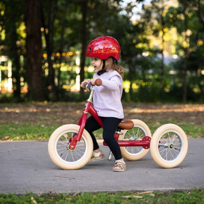 Child riding a red Trybike balance bike with trike mode on a paved path in a park.