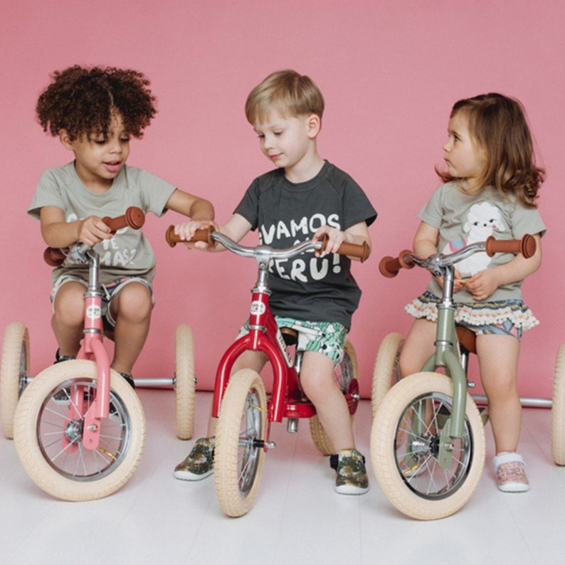 Three children riding balance bikes against a pink background